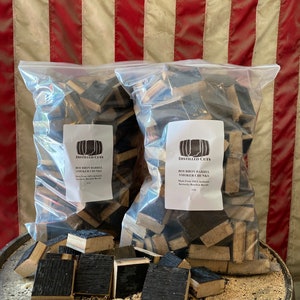 May include: Two clear plastic bags filled with dark brown bourbon barrel smoker chunks. The bags are labeled "DISTILLED CUTS BOURBON BARREL SMOKER CHUNKS" and are sitting on a wooden barrel with sawdust.