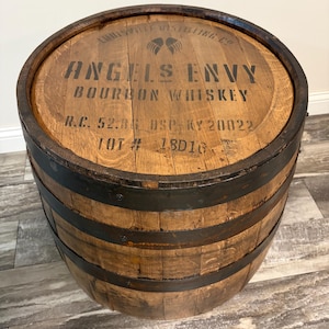 May include: A cylindrical wooden bourbon barrel with dark metal hoops. The top surface features the black printed text "LOUISVILLE DISTILLING CO", "ANGELS ENVY", "BOURBON WHISKEY", "R.C. 52.86", "DSP KY 20022", and "LOT # 18D16" with a wing icon.