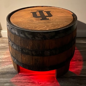 May include: A round wooden barrel table featuring a dark brown finish with black metal hoops and rivets. The top surface displays a black Indiana University IU logo. The base of the barrel is illuminated by a bright red light glowing from underneath.