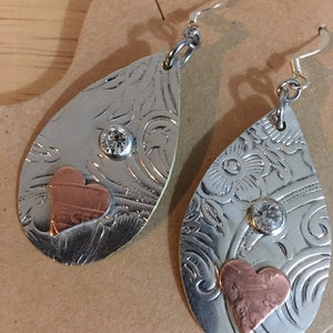 May include: A pair of silver teardrop-shaped earrings with a copper heart and a clear rhinestone on each earring.