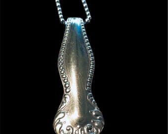 May include: A silver spoon pendant necklace with a decorative design at the bottom. The spoon is attached to a silver chain.