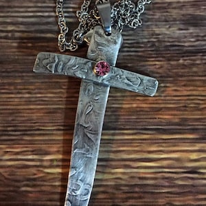 May include: A silver cross pendant with a pink rhinestone in the center. The cross has a textured, hammered finish and is attached to a silver chain.