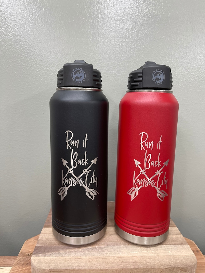 May include: Two stainless steel water bottles with black and red powder coated finishes. Both bottles have a silver lid with a logo that says "Polar Camel". The bottles have the text "Run it Back Kansas City" with crossed arrows engraved on the front.