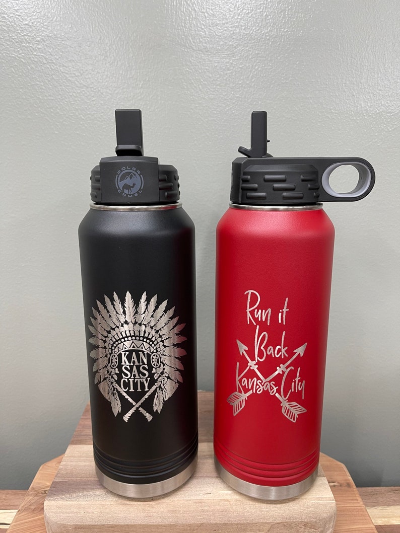 May include: Two stainless steel water bottles with lids and handles. The black bottle has a Native American headdress design with the text "Kansas City". The red bottle has the text "Run it Back Kansas City" with two crossed arrows.