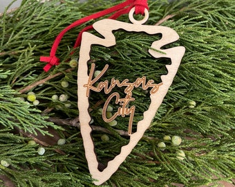 Op de afbeelding: Een houten pijlpunt ornament met de woorden "Kansas City" in cursief erop geschreven. Het ornament hangt aan een rood lint en is omringd door groene dennentakken.