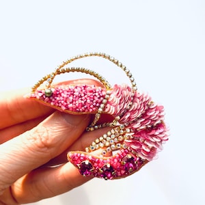 May include: A handcrafted, pink beaded brooch in the shape of a shrimp. The brooch features a gold-colored wire frame, embellished with sequins, beads, and rhinestones. The shrimp's body is a gradient of pink hues, with a curved design.