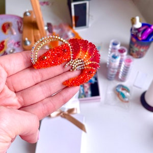 May include: A red and gold beaded lobster brooch. The lobster is made of small red beads and has gold accents. The brooch is a unique and eye-catching piece of jewelry.