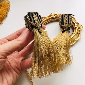 May include: A pair of gold tassel earrings with black and gold beaded embellishments. The earrings are dangling and have a long tassel.