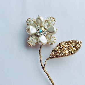 May include: A handcrafted floral brooch featuring a flower with white pearl petals and a central blue crystal. The stem and leaf are detailed with gold beads and wire, creating a delicate, elegant accessory.
