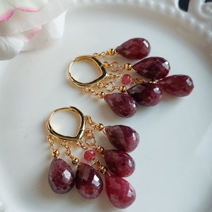 Peut inclure: Une paire de boucles d'oreilles dorées avec de multiples pierres précieuses rouges profondes en forme de goutte d'eau facettées et pendantes. Les boucles d'oreilles sont présentées sur une assiette blanche décorative.