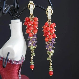 May include: A pair of long, dangling earrings with a gold chain and red, purple, and green beads. The earrings are on a red velvet mannequin bust.
