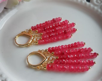 Puede incluir: Un par de pendientes colgantes con aros dorados y múltiples hebras de cuentas facetadas de color rojo brillante. Los pendientes se exhiben sobre un plato blanco decorativo, con una flor rosa y blanca en el fondo.
