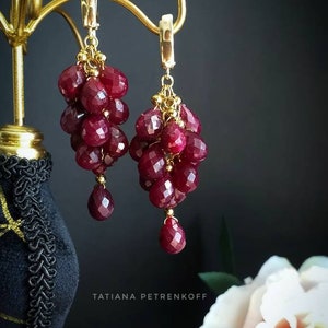 Peut inclure: Une paire de boucles d'oreilles en or avec des grappes de perles de pierres précieuses rouge foncé qui pendent. Les boucles d'oreilles sont présentées sur un support en or sur un fond noir.
