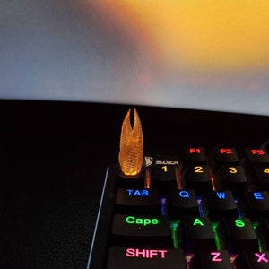 May include: A clear, translucent, amber-coloured keycap with a textured, geometric design. The keycap is shaped like a tall, pointed spire. It is sitting on a black keyboard with rainbow-coloured backlit keys.