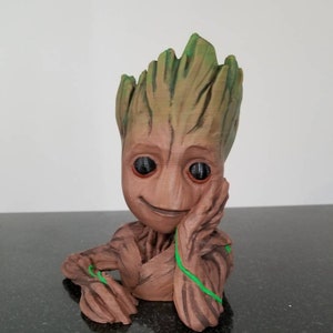 Puede incluir: Una figura impresa en 3D de color marrón y verde de un personaje de la película Guardianes de la Galaxia. El personaje está sentado con los brazos cruzados y tiene una vid verde envuelta alrededor de sus brazos. La figura tiene una cara sonriente con ojos grandes.