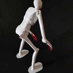 May include: A white, skeletal figurine with red accents on the hands and neck, posed in a crouching position. The figure is standing on two white, oval-shaped bases against a black background. The figurine has long arms and legs.