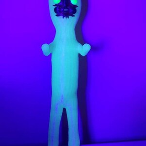 Puede incluir: Una mu&ntilde;eca verde que brilla en la oscuridad con una cara en blanco y negro. La mu&ntilde;eca tiene un dise&ntilde;o simple con una cabeza redonda, dos brazos y dos piernas.