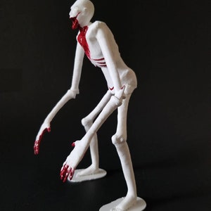 May include: A white, skeletal figurine with red accents. The figure has long limbs and a contorted pose, with red paint detailing the mouth, chest, and hands. The figurine stands on a white base against a dark background.