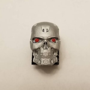 May include: A silver-colored robotic skull figurine featuring a textured metallic surface and two circular, bright red eyes. The skull has a detailed mechanical jaw and facial structure, mounted on a small black base, presenting a front-facing view.