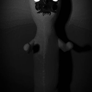 Puede incluir: Una figura en blanco y negro con una cabeza puntiaguda y grandes ojos blancos. La figura tiene una boca con dientes y parece estar hecha de papel mach&eacute;.