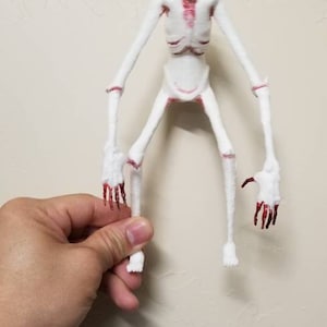 May include: A white, skeletal figurine with red accents on the chest, face, and hands. The figure has long, thin limbs and is held against a textured, off-white wall. The figurine is a unique, possibly horror-themed collectible.