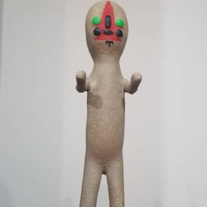 Puede incluir: Una figurita de color beige de una criatura humanoide con una cara triangular roja y ojos verdes. La figurita tiene un dise&ntilde;o simple y est&aacute; de pie con los brazos extendidos.