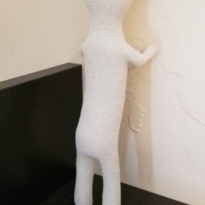 Puede incluir: Una figurita humanoide blanca texturizada con una cabeza redonda y miembros largos y delgados. La figurita est&aacute; de pie en un estante negro y apoyada contra una pared blanca.