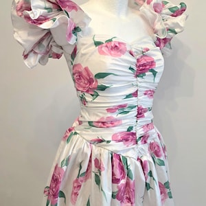 May include: A white dress with a pink rose floral pattern. The dress features ruffled sleeves and a ruched bodice. The skirt is gathered at the waist. The dress is displayed on a mannequin.