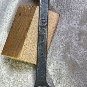 May include: A vintage metal wrench with open ends, marked with "9/16" and "1/2". The wrench has the text "44 CORNWELL EW 18" on the handle. It rests on a small piece of wood, set against a textured, light-colored surface.