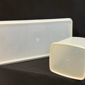 May include: Two off-white Tupperware containers. One is rectangular with a lid, the other is a square container. Both have rounded corners and are made of translucent plastic. The lid is rectangular and has a slightly raised edge.