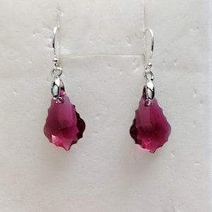 May include: A pair of dangle earrings featuring faceted, teardrop-shaped, deep magenta crystals. The crystals are suspended from silver-colored hooks. The earrings are displayed against a white background.
