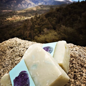 May include: Two bars of white soap with a blue and purple label. The label has a wax seal with a design. The soap is sitting on a rock with a mountain range in the background.