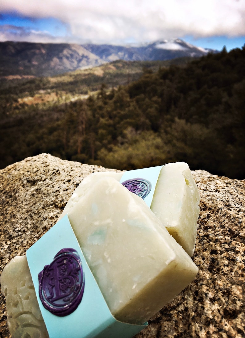 May include: Two bars of white soap with a blue and purple label. The label has a wax seal with a design. The soap is sitting on a rock with a mountain range in the background.