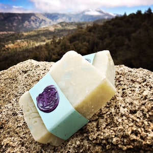 May include: Two bars of white soap with a light blue label and a purple wax seal. The soap is sitting on a rock with a mountain range in the background.