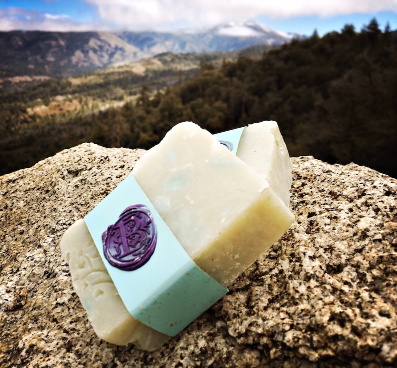 May include: Two bars of white soap with a light blue label and a purple wax seal. The soap is sitting on a rock with a mountain range in the background.
