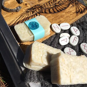 May include: Two bars of handmade soap, one with a tree design and the other with a rune symbol. The soap is on a wooden board with a carved raven design. There are also several white stones with runes carved on them.