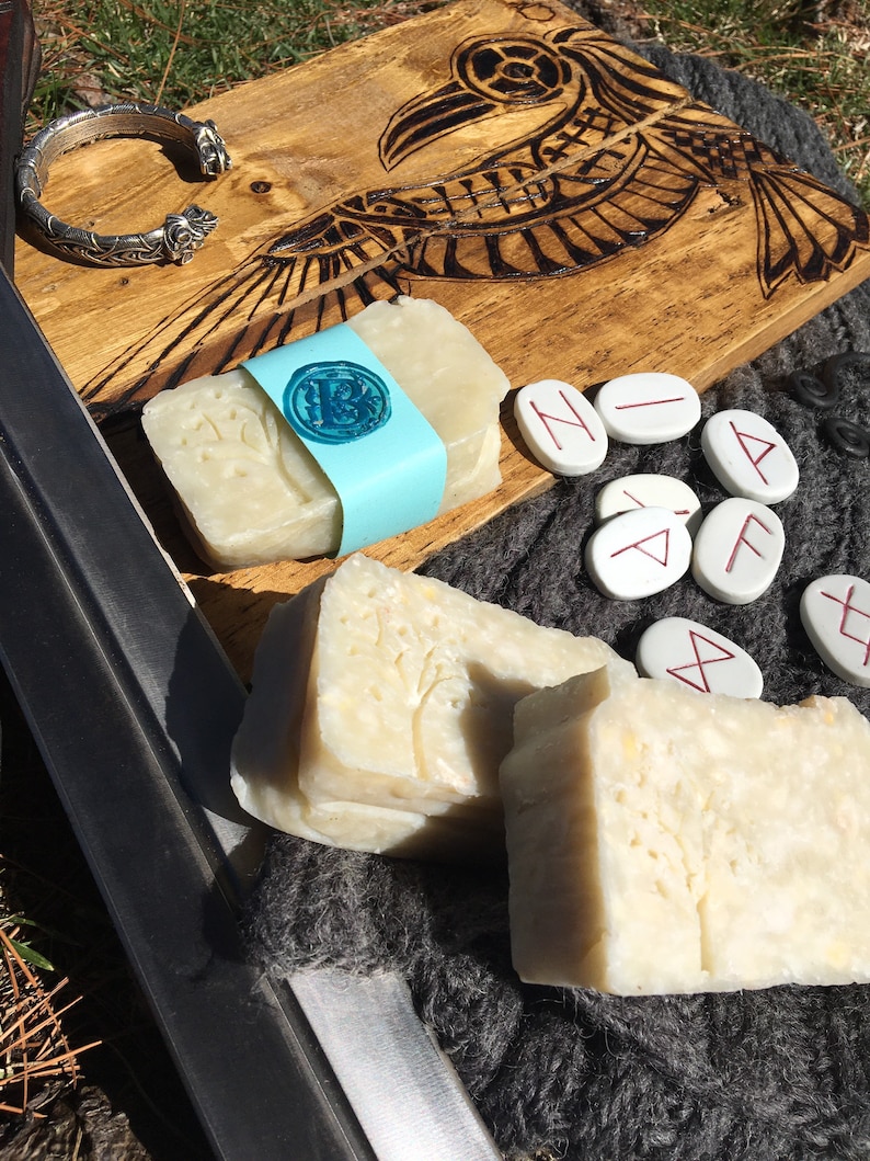 May include: Two bars of handmade soap, one with a tree design and the other with a rune symbol. The soap is on a wooden board with a carved raven design. There are also several white stones with runes carved on them.