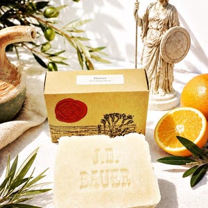 Peut inclure: Un pain de savon rectangulaire blanc cassé portant l'inscription en relief J.D. BAUER, placé devant une boîte en carton beige avec un sceau en cire rouge et une étiquette indiquant Athena The Clarity Ritual, accompagné d'une orange et d'olivier.