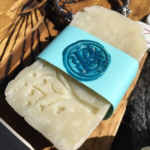 May include: A white bar of soap with a floral design wrapped in a light blue ribbon with a blue wax seal. The seal has a letter "B" in the center.