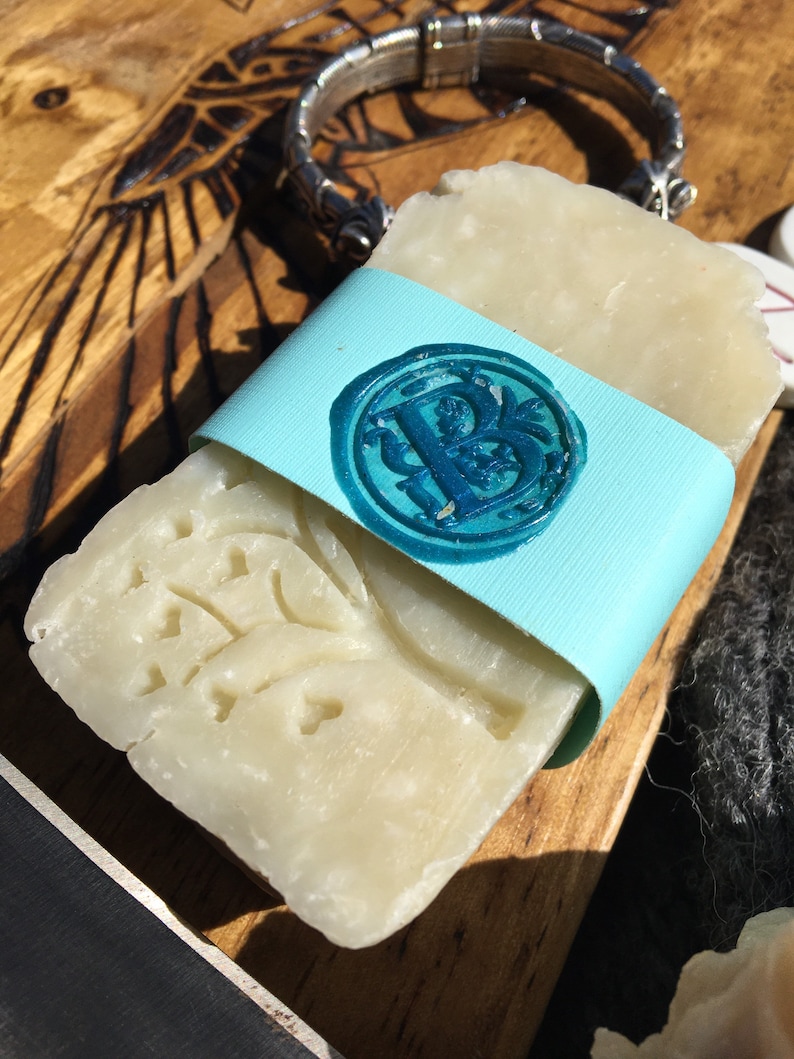 May include: A white bar of soap with a floral design wrapped in a light blue ribbon with a blue wax seal. The seal has a letter "B" in the center.