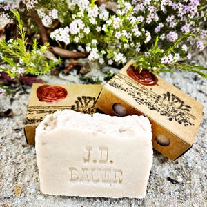 May include: A bar of natural soap with the words "J.D. Bauer" embossed on the top. The soap is sitting on a sandy surface with two brown boxes behind it. The boxes have a red wax seal and a design of a tree on them.
