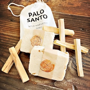 May include: A white cloth bag with the text "Palo Santo" and "Wild Harvested Peru" printed on it. The bag contains several sticks of Palo Santo wood, which are used for smudging and aromatherapy. The sticks are arranged in a cross shape and have a natural brown color.