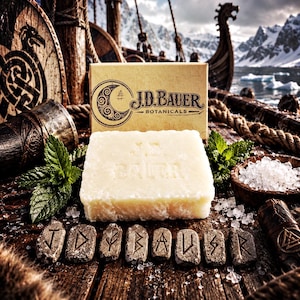 May include: A rectangular off-white bar of soap with the embossed text J.D. BAUER, positioned in front of a branded cardboard box. The soap is surrounded by fresh green mint leaves, coarse white salt crystals, and seven stone runes on wood.