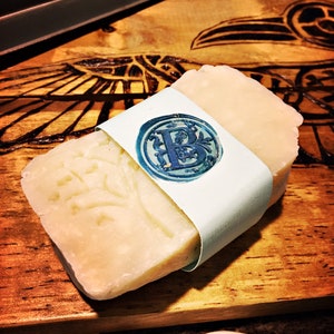 May include: A bar of white soap with a decorative design wrapped in a light blue paper with a wax seal featuring the letter "B".