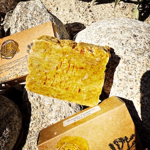 Może przedstawiać: Złotożółta kostka mydła z wytłoczonym napisem "BAHIA" spoczywa na skałach. Widoczne jest również małe, brązowe kartonowe pudełko ze złotą pieczęcią i napisem "Orange, Turmeric & Tea Tree". Naturalny produkt do pielęgnacji skóry.