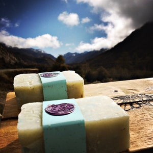 May include: Two bars of natural soap with a light blue and white label. The label has a purple wax seal with a design. The soap is sitting on a wooden surface with a mountain range in the background.