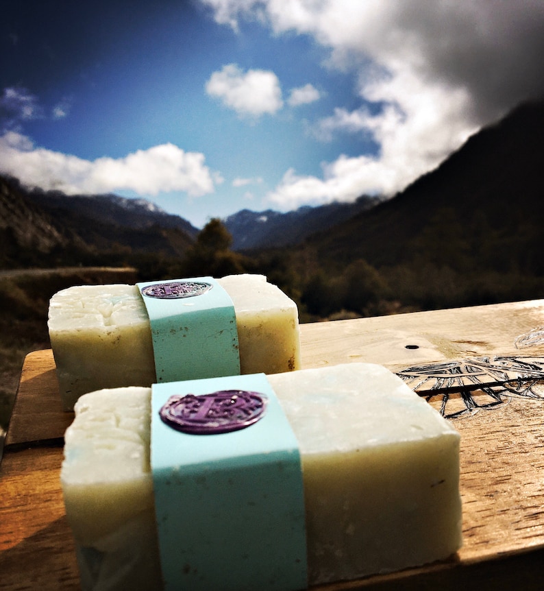 May include: Two bars of natural soap with a light blue and white label. The label has a purple wax seal with a design. The soap is sitting on a wooden surface with a mountain range in the background.