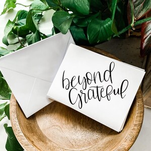May include: A white card with black calligraphy that reads "beyond grateful" on a wooden surface.