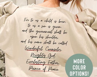 May include: Cream-coloured sweatshirt with black script text: "For to us a child is born..." and "MORE COLOR OPTIONS!" in a green circle. The text is accented with red and green lines. A casual, comfortable top.
