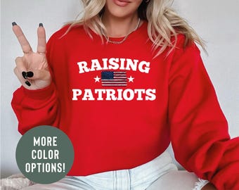 May include: A red sweatshirt with the words "RAISING PATRIOTS" in white block letters, with an American flag graphic. The sweatshirt also has a circular graphic that says "MORE COLOR OPTIONS!"